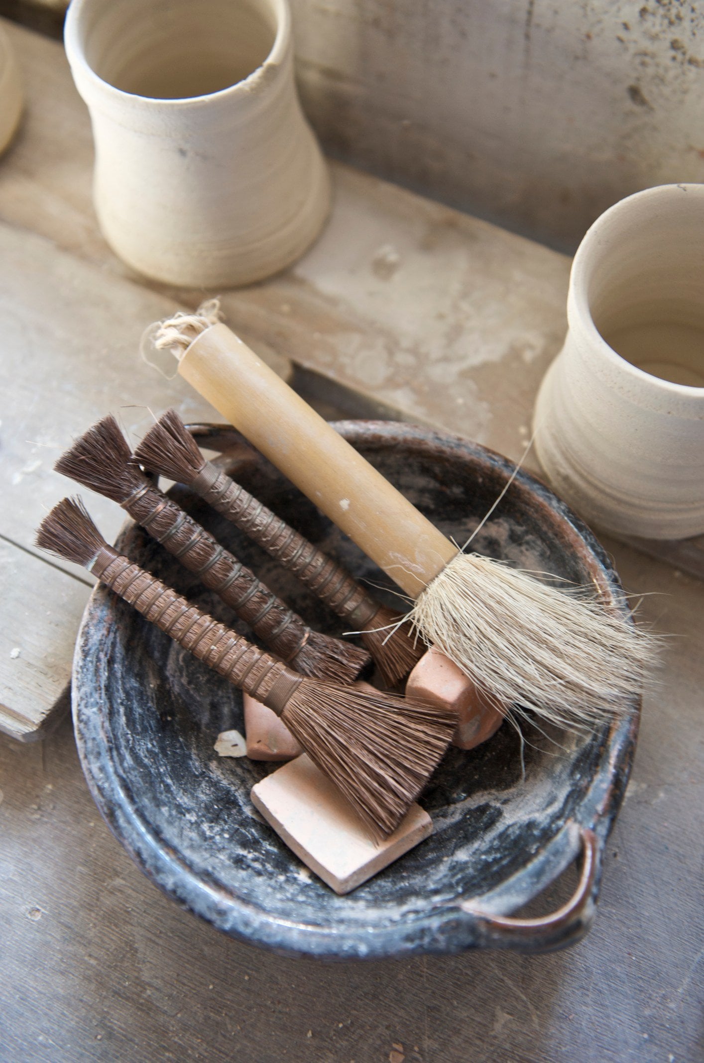 Bundle of hair and fibre brushes in a dark glazed bowl on a studio shelf, with two unglazed clay cups behind.