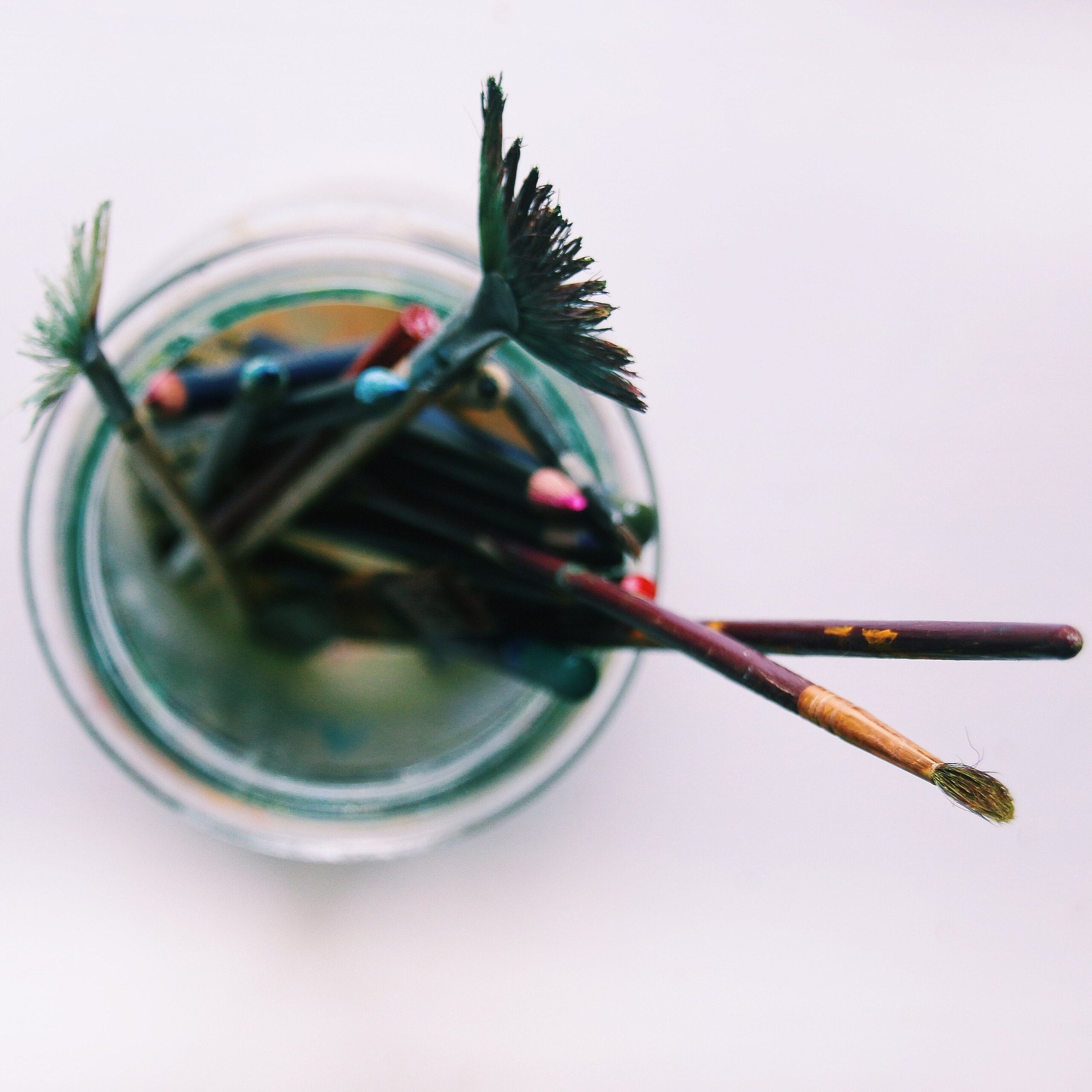 Top-down view of a glass jar holding used paintbrushes and pencils on a pale lilac-white surface, with two brushes resting across the rim.