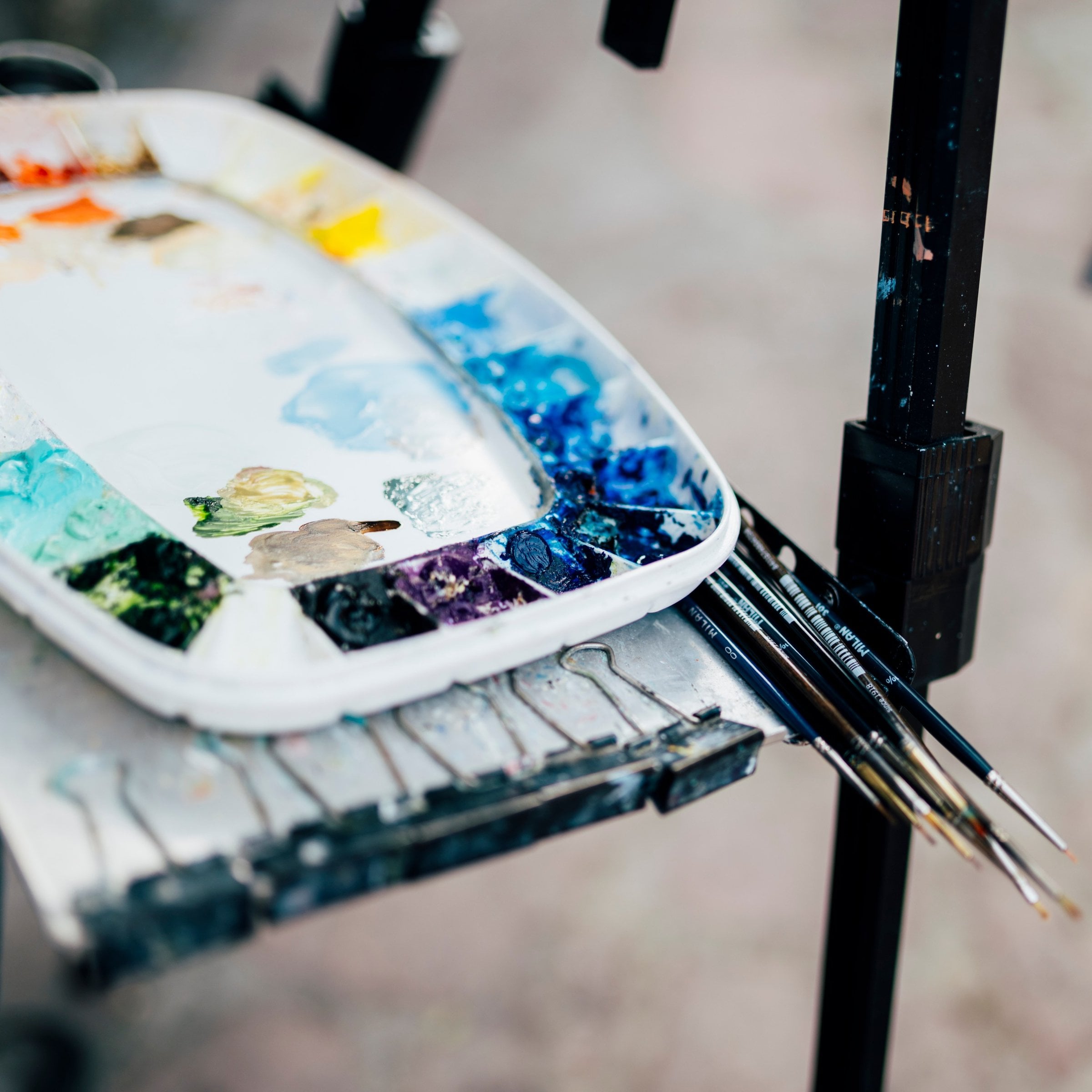 White mixing palette loaded with blue, teal, yellow, orange and red oil paint, resting on an easel tray with fine brushes tucked beside it.