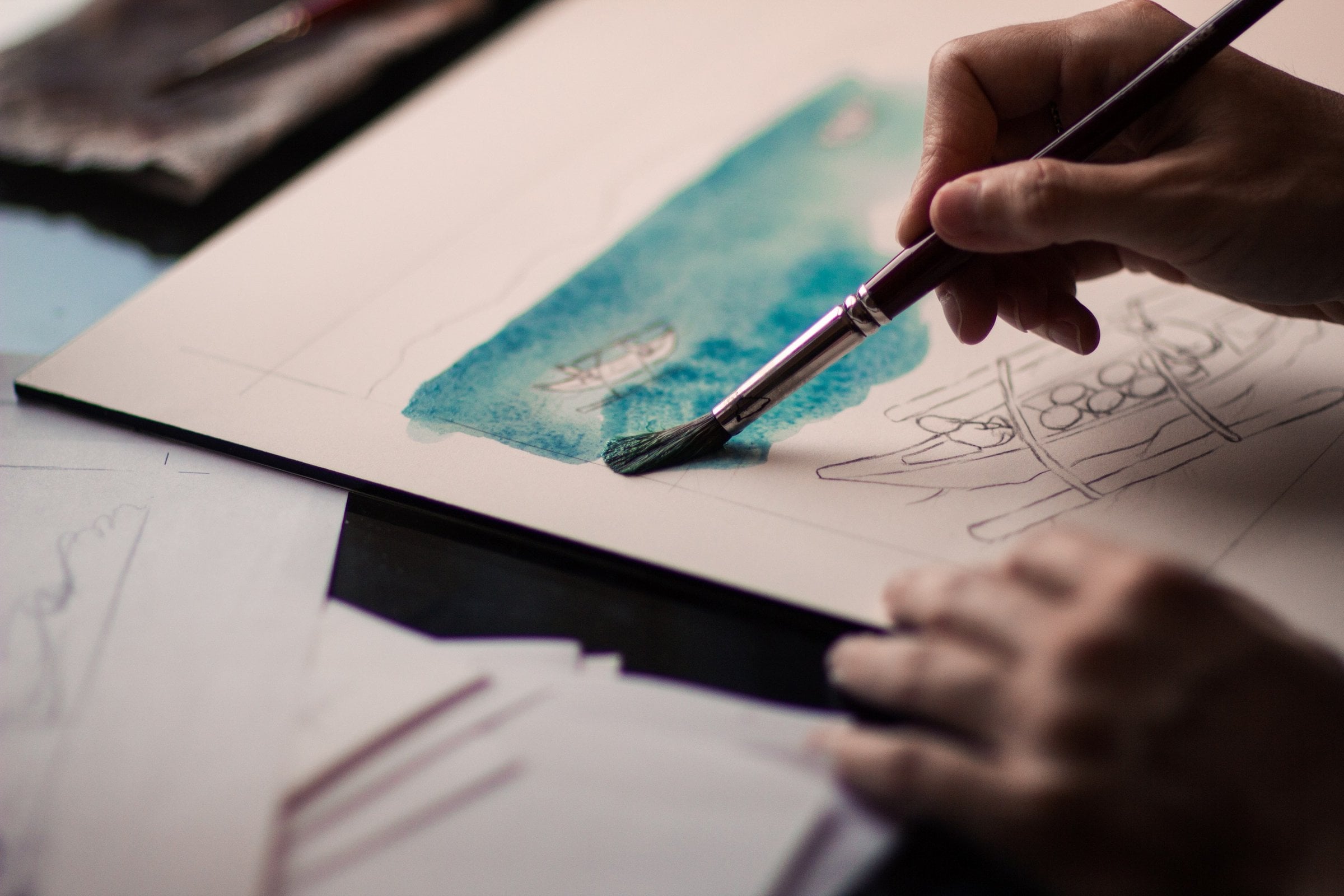 Hand painting a teal watercolour wash onto pencil-sketched paper on a drawing board while the other hand steadies the sheet.