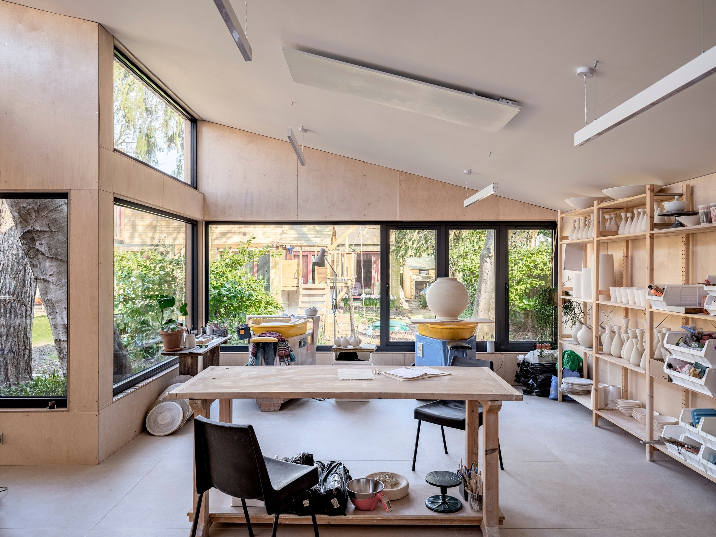 Daylight pottery studio with pale plywood walls, large windows onto a garden, a wooden worktable with two black chairs and open shelving of white ceramic vases and bowls.