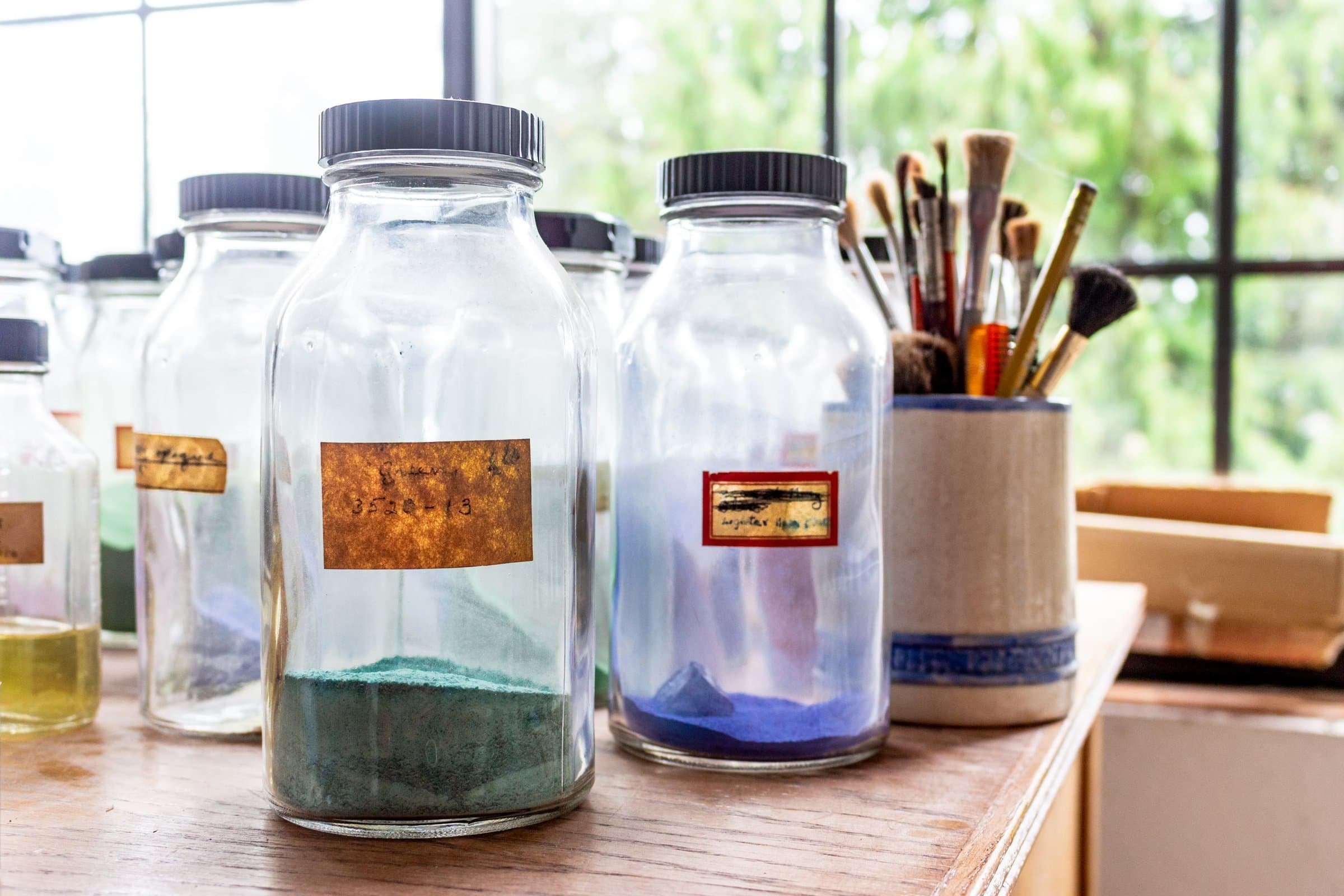 Glass jars of green and blue powdered pigment with handwritten labels beside a ceramic pot of brushes, on a wooden table in front of a leafy window.