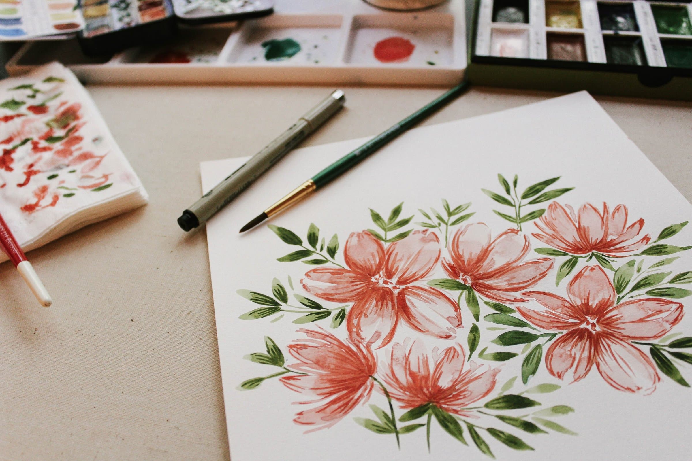 Watercolour painting of coral-red flowers with green leaves on paper, beside a fine-liner pen, a round brush and open paint palettes on a cream cloth.