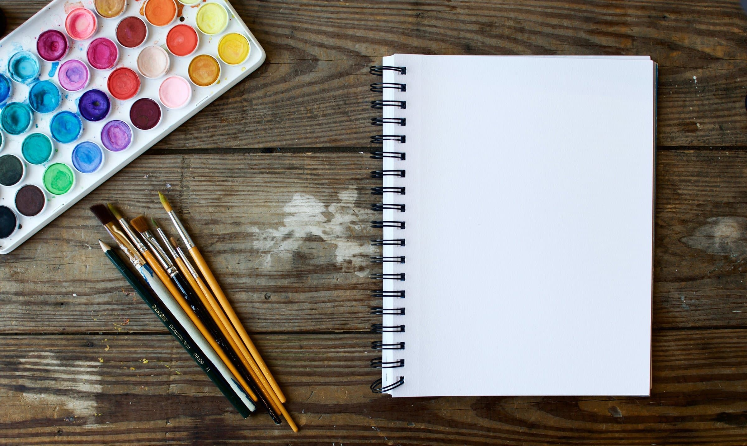 Overhead view of a round-pan watercolour set, a handful of brushes and a blank spiral sketchbook on a weathered wooden table.